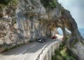 Two sports cars, including a striking Ferrari from the Cavalcade Siviglia, drive along a winding mountain road in Andalusia, framed by a rocky archway and steep cliffs on one side.