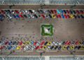 Aerial view of many parked cars, including Ferraris, in rows on both sides of a square with a statue in the center in Andalusia, as people walk around during the Cavalcade Siviglia event.
