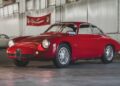A classic red sports car from the RM Sotheby’s Quadrifoglio Collection is parked indoors on a concrete floor, with white brick walls and large windows in the background.