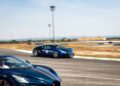 A blue Bugatti sports car speeds on a racetrack in Portugal, with another dark-colored Bugatti in the foreground. The track is surrounded by dry grass and distant buildings, capturing the thrill of the Grand Tour 2025.