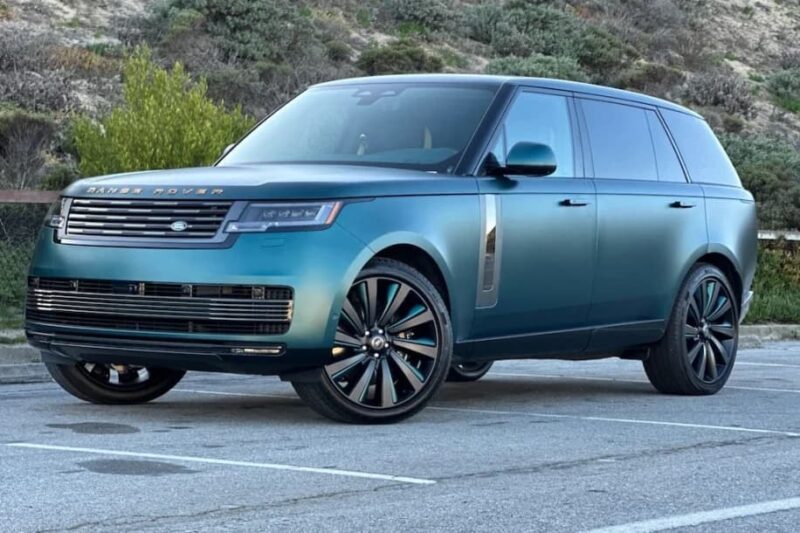 A metallic blue-green Range Rover SV Carmel Edition SUV is parked in a lot with trees and shrubs in the background, highlighting the exclusivity of this sought-after model among Rovers for sale.
