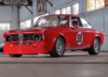 A red vintage race car with the number 99 on the door is parked inside an industrial-style garage with concrete floors and large windows, ready for display at the RM Sotheby’s Quadrifoglio Collection Monterey 2025.