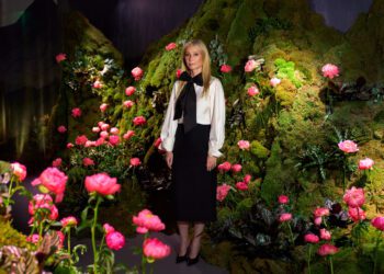 A woman stands in front of a green, mossy backdrop decorated with pink flowers at Genesis House’s The Forest Within, wearing a black skirt, white blouse, and black shoes—evoking Gwyneth Paltrow’s effortless elegance.