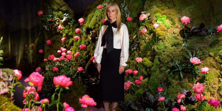 A woman stands in front of a green, mossy backdrop decorated with pink flowers at Genesis House’s The Forest Within, wearing a black skirt, white blouse, and black shoes—evoking Gwyneth Paltrow’s effortless elegance.