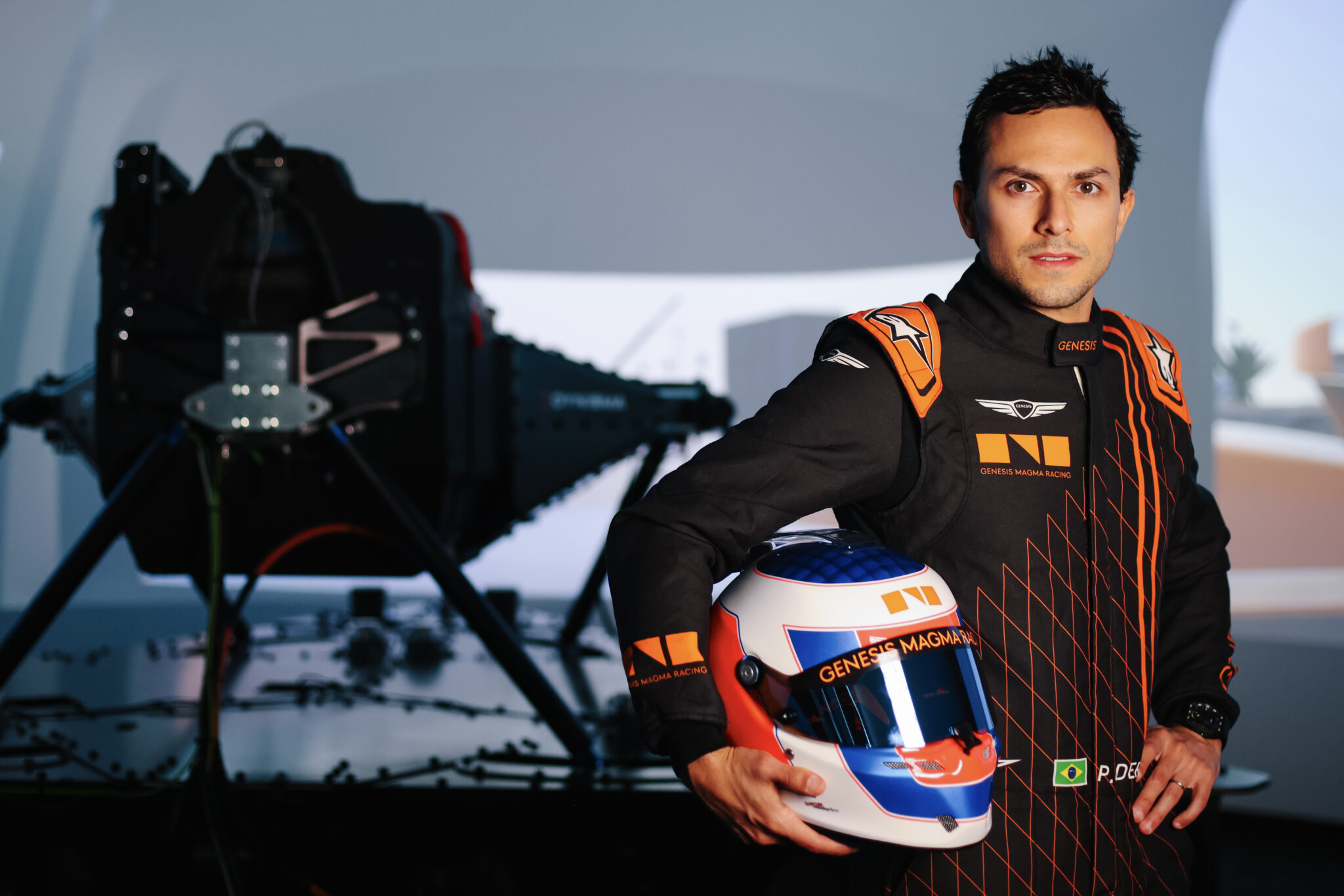A racing driver in a black and orange suit stands indoors holding a helmet, with Genesis Magma Racing gear and the GMR-001 Hypercar Simulator for virtual racing visible in the blurred background.