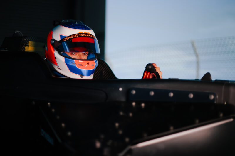 A race car driver wearing a helmet sits in the cockpit of a Genesis Magma Racing GMR-001 Hypercar Simulator, gripping the steering wheel, with a blurred fence in the background.