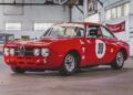 A red vintage Alfa Romeo race car with the number 99 on the door is parked indoors in a garage with white walls and large windows, part of the RM Sotheby’s Quadrifoglio Collection Monterey 2025.