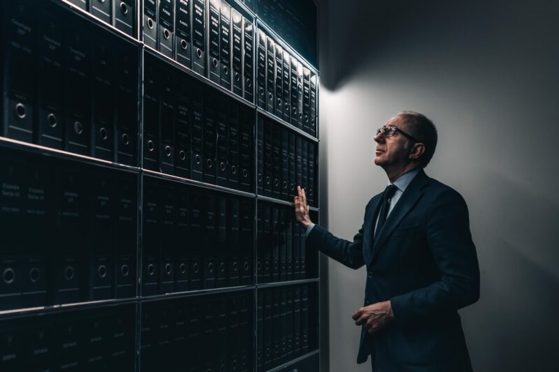 A man in a suit stands in a dimly lit room, reaching toward shelves filled with black binders organized in rows—each binder chronicling Lamborghini’s rich automotive history for Polo Storico.
