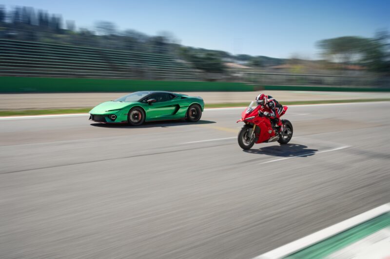 A Lamborghini Temerario and a Ducati Panigale V4 race head-to-head on the track, their green and red colors streaked with motion blur that showcases their incredible speed.