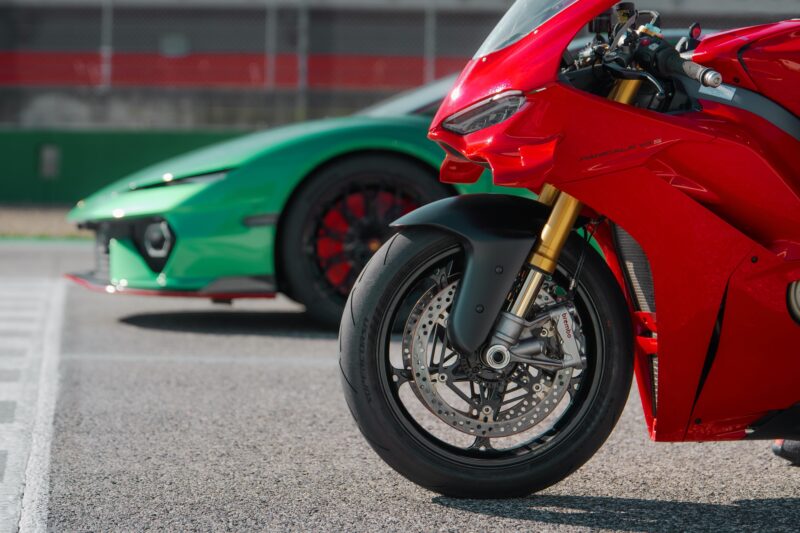 A red Ducati Panigale V4 and a green Lamborghini Temerario are parked side by side on a racetrack, their head-to-head rivalry highlighted by a focus on the motorcycle's striking front wheel.