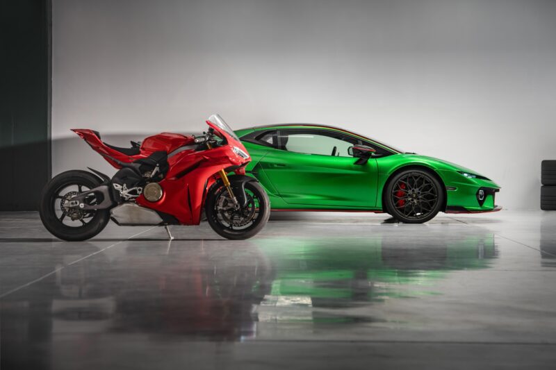 A red Ducati Panigale V4 and a green Lamborghini Temerario stand head-to-head in a clean, modern indoor garage with a reflective floor.