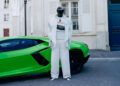 A person wearing a black helmet and all-white Balenciaga outfit stands in front of a bright green Lamborghini on a city street.