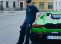 A person in dark casual Balenciaga clothing and a visor leans against a green Lamborghini parked on a quiet city street.