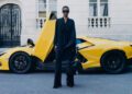 A person in black Balenciaga clothing and sunglasses stands in front of a yellow Lamborghini with its door open on a city street.
