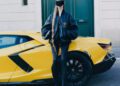 Person in black Balenciaga jacket, tights, and visor stands in front of a yellow Lamborghini parked on a city street, with green double doors in the background.