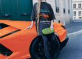 A woman in oversized Lamborghini x Balenciaga collaboration clothing and a large visor stands next to an orange Lamborghini sports car on a city street.