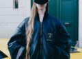 A person with long hair wears a large black leather jacket from the Balenciaga x Lamborghini collaboration and black visor sunglasses, standing in front of a yellow sports car and green door.