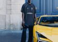 A man in black Lamborghini-branded clothing and cap stands next to a yellow sports car, highlighting a possible Lamborghini-Balenciaga collaboration, in front of a blue door and stone building.