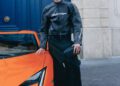 A person in black Balenciaga clothing and a visor stands next to an orange Lamborghini sports car in front of a blue door and beige stone building.