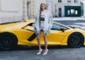 A woman in a silver fringed jacket and white heels stands in front of a yellow Lamborghini parked on a city street.