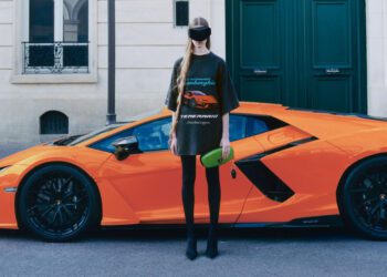 A person wearing black Balenciaga clothing, a visor, and holding a green clutch stands in front of an orange Lamborghini parked on a city street.