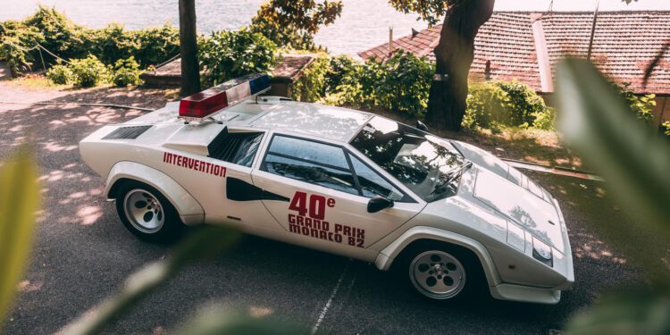 White Lamborghini Countach classic with "40e Grand Prix Monaco 82" and "Intervention" markings, parked outdoors near trees and a body of water, featuring a rooftop light bar—a true icon among race cars.