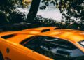 Close-up of the rear section of a yellow Lamborghini sports car with a rear wing, parked outdoors near trees and a body of water.