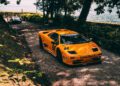 A classic yellow Lamborghini sports car is parked on a gravel pathway near a body of water, with another car visible in the background, surrounded by trees and greenery.