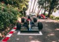 A black race car with the number 34 is parked on a road lined with trees and greenery, with two other classic race cars visible in the background.