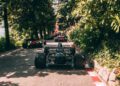 A row of classic formula race cars is parked on a narrow, tree-lined road bordered by greenery and red-and-white curbs.