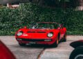 A red classic sports car with pop-up headlights is parked on a driveway, surrounded by greenery and trees in the background—a true favorite among race car enthusiasts and lovers of classics.