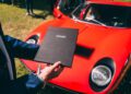 A person holds a black Lamborghini folder in front of a red classic Lamborghini sports car, showcasing one of the timeless race car classics parked on the grass.