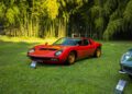 A red vintage Lamborghini sports car with gold wheels is displayed on grass, surrounded by green trees; informational signs are placed in front of this classic.