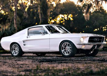 A classic white Ghost Mustang Fastback parked outdoors on a dirt surface, with trees and a wooden fence in the background—a nod to modern muscle and Roush Performance heritage.