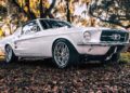 A white classic Ford Mustang, inspired by ECD’s Bespoke '67 Ghost Mustang Fastback and powered by Roush Performance, is parked outdoors on fallen leaves, with trees and sunlight adding to its timeless allure.