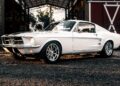 A white ECD’s Bespoke '67 Ghost Mustang Fastback parked on gravel in front of a barn, sunlight reflecting off its polished chrome wheels—now boasting modern muscle thanks to Roush Performance.