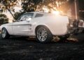 ECD’s Bespoke '67 Ghost Mustang Fastback with chrome wheels is parked outdoors on gravel at sunset, boasting modern muscle from Roush Performance, framed by trees and golden sunlight in the background.