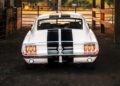 A classic white ECD’s Bespoke '67 Ghost Mustang Fastback with black racing stripes is parked in a barn, viewed from the rear with its taillights on, showcasing a dose of modern muscle from Roush Performance.