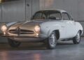 A classic white Alfa Romeo coupe, part of the RM Sotheby’s Quadrifoglio Collection, is parked indoors with its headlights on, set against a backdrop of corrugated metal and brick walls.