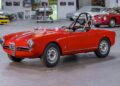 A classic red Alfa Romeo convertible with a roll bar, part of the RM Sotheby’s Quadrifoglio Collection Monterey 2025, is parked indoors among other vintage cars in a spacious garage.