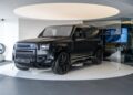 A matte black Land Rover Defender SUV, perfect for a unique garage creation, is displayed on a circular white platform inside a modern showroom with dark and light decor elements.