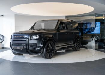 A matte black Land Rover Defender SUV, perfect for a unique garage creation, is displayed on a circular white platform inside a modern showroom with dark and light decor elements.