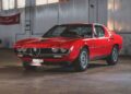 A classic red Alfa Romeo from the RM Sotheby’s Quadrifoglio Collection is parked indoors on a concrete floor near a white brick wall and large windows, capturing the elegance ahead of Monterey 2025.