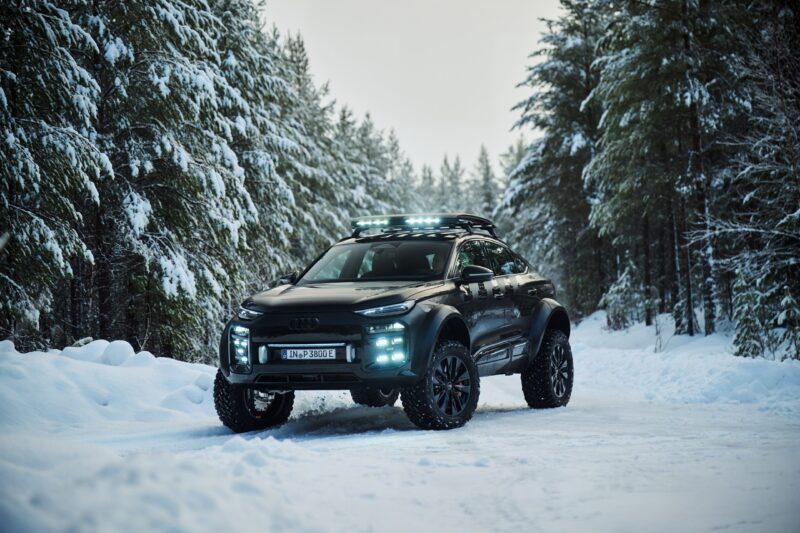 A black Audi SUV with off-road tires and roof lights embodies Expedition Elegance as it’s parked on a snowy forest road, surrounded by snow-covered trees.