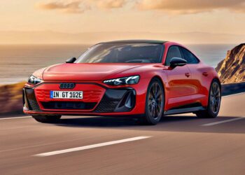 A red Audi electric sports car, inspired by the Most Affordable Audi e-tron GT Debuts And Its Anything But Basic, drives along a coastal road with cliffs and the ocean visible in the background under a partly cloudy sky.