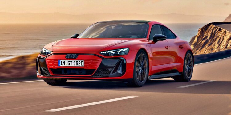 A red Audi electric sports car, inspired by the Most Affordable Audi e-tron GT Debuts And Its Anything But Basic, drives along a coastal road with cliffs and the ocean visible in the background under a partly cloudy sky.