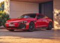 An Audi e-tron GT in striking red is parked in a driveway, plugged into a wall-mounted charging station beside a modern house, showcasing the debut of affordable luxury electric vehicles.