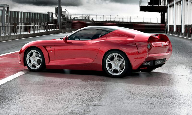 A red sports car, reminiscent of the significance of the 1-of-1 Alfa Romeo DoppiaCoda by Zagato, is parked on a wet racetrack near a building under an overcast sky.