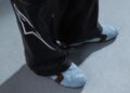 Person wearing black pants with white design and light blue socks with brown accents, standing on a light gray carpeted floor, completing their look with STARLINE Sneaker inspired by F1 DNA.