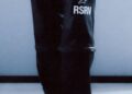Person wearing baggy black pants with "RSRV" and a logo on the left leg, paired with STARLINE Sneakers inspired by F1 DNA, standing on a light-colored floor, partially visible from the knees down.